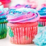 cotton candy cupcakes decorated with pink and blue frosting and topped with cotton candy ready to be enjoyed. Bits of cotton candy are scattered around the cupcakes as well.