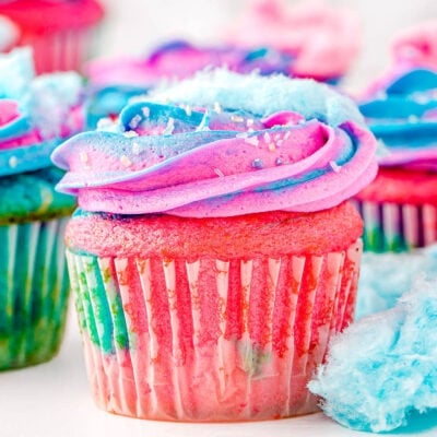 cotton candy cupcakes decorated with pink and blue frosting and topped with cotton candy ready to be enjoyed. Bits of cotton candy are scattered around the cupcakes as well.