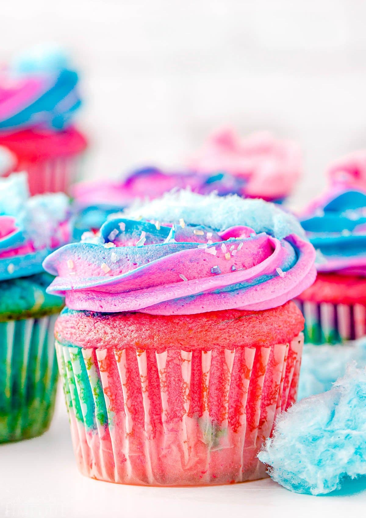 lots of cotton candy cupcakes decorated with pink and blue frosting and topped with cotton candy ready to be enjoyed. Bits of cotton candy are scattered around the cupcakes as well.