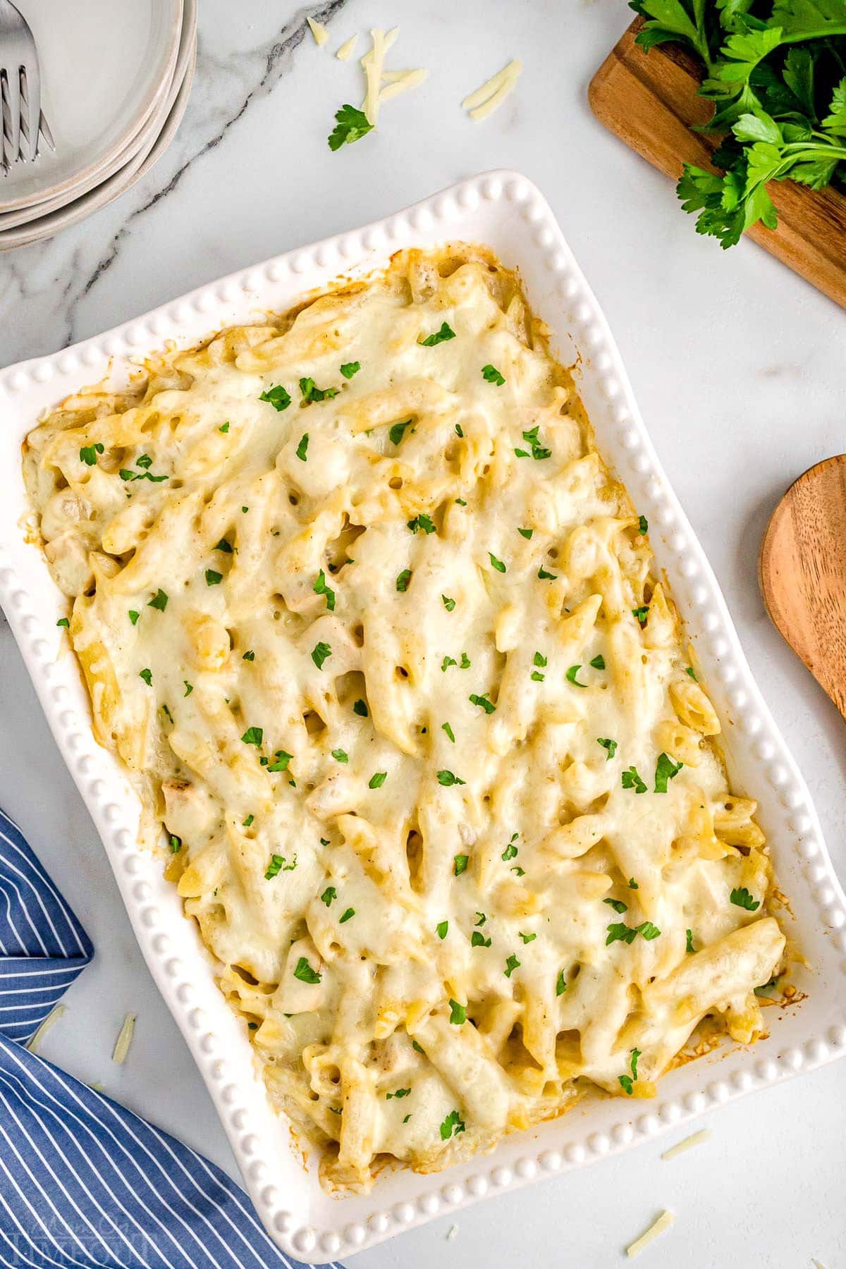 top down look at chicken alfredo bake in a white casserole dish garnished with fresh parsley and blue and white striped napkin in the bottom left corner.