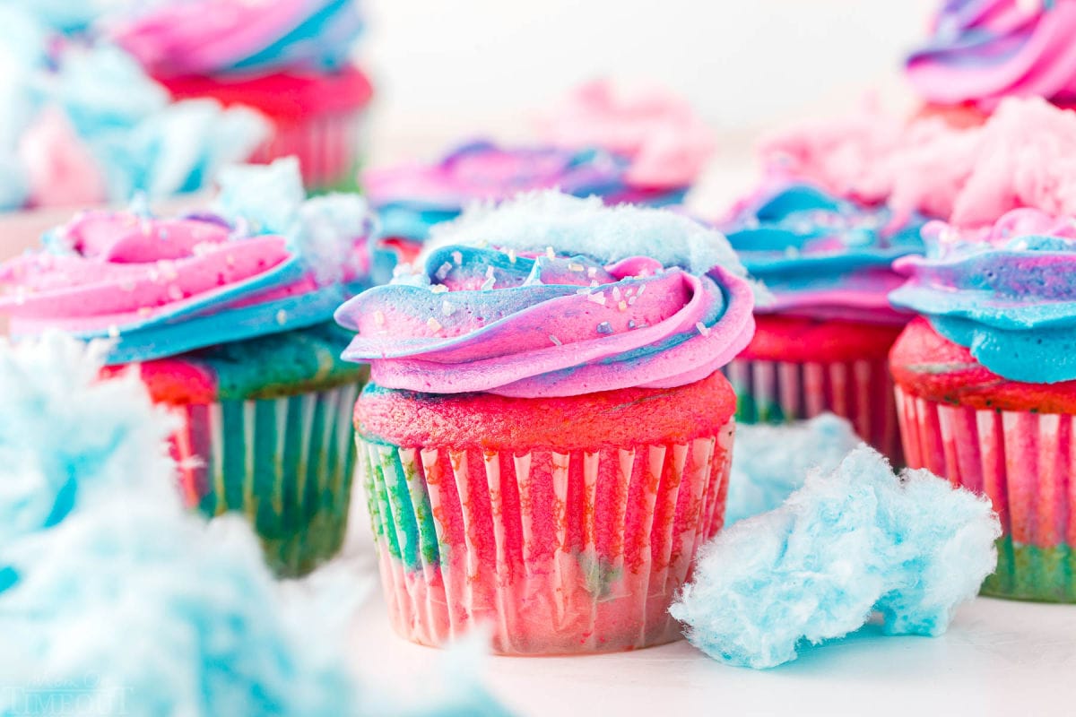 lots of cotton candy cupcakes decorated with pink and blue frosting and topped with cotton candy ready to be enjoyed. Bits of cotton candy are scattered around the cupcakes as well.
