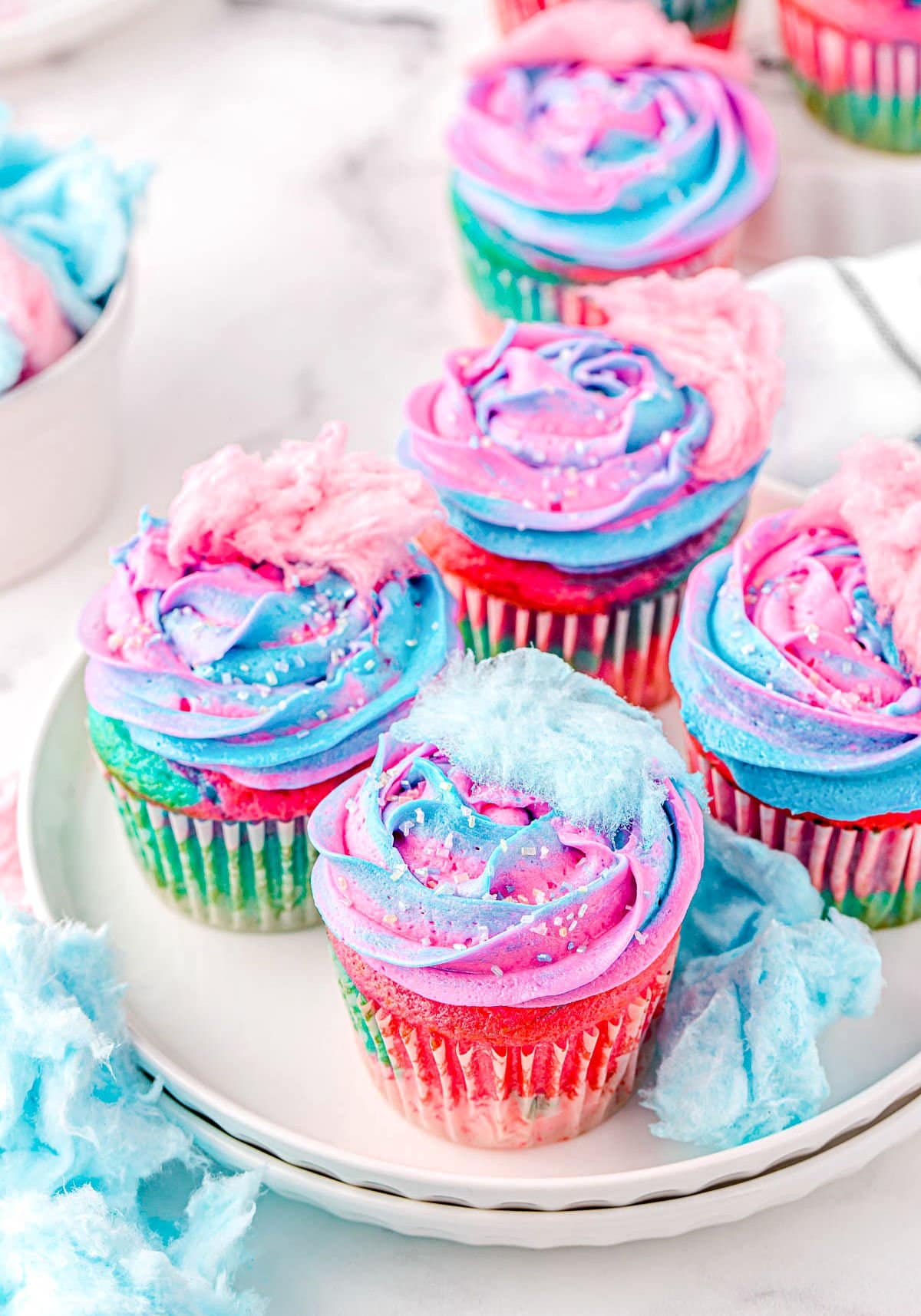 five cotton candy cupcakes sitting on a white plate topped with swirled frosting and small pieces of cotton candy.