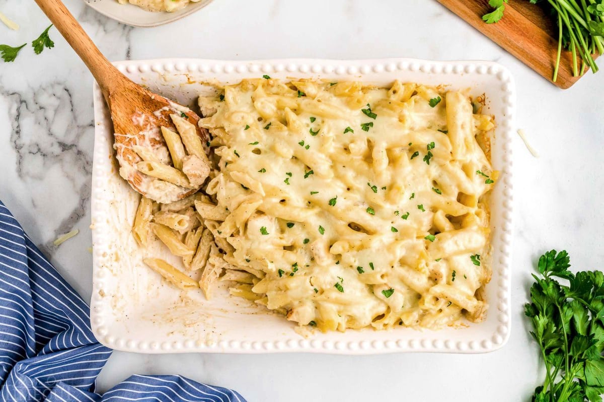 top down view of white casserole baking dish with some of the chicken alfredo bake already served. wood spoon is left in the dish.