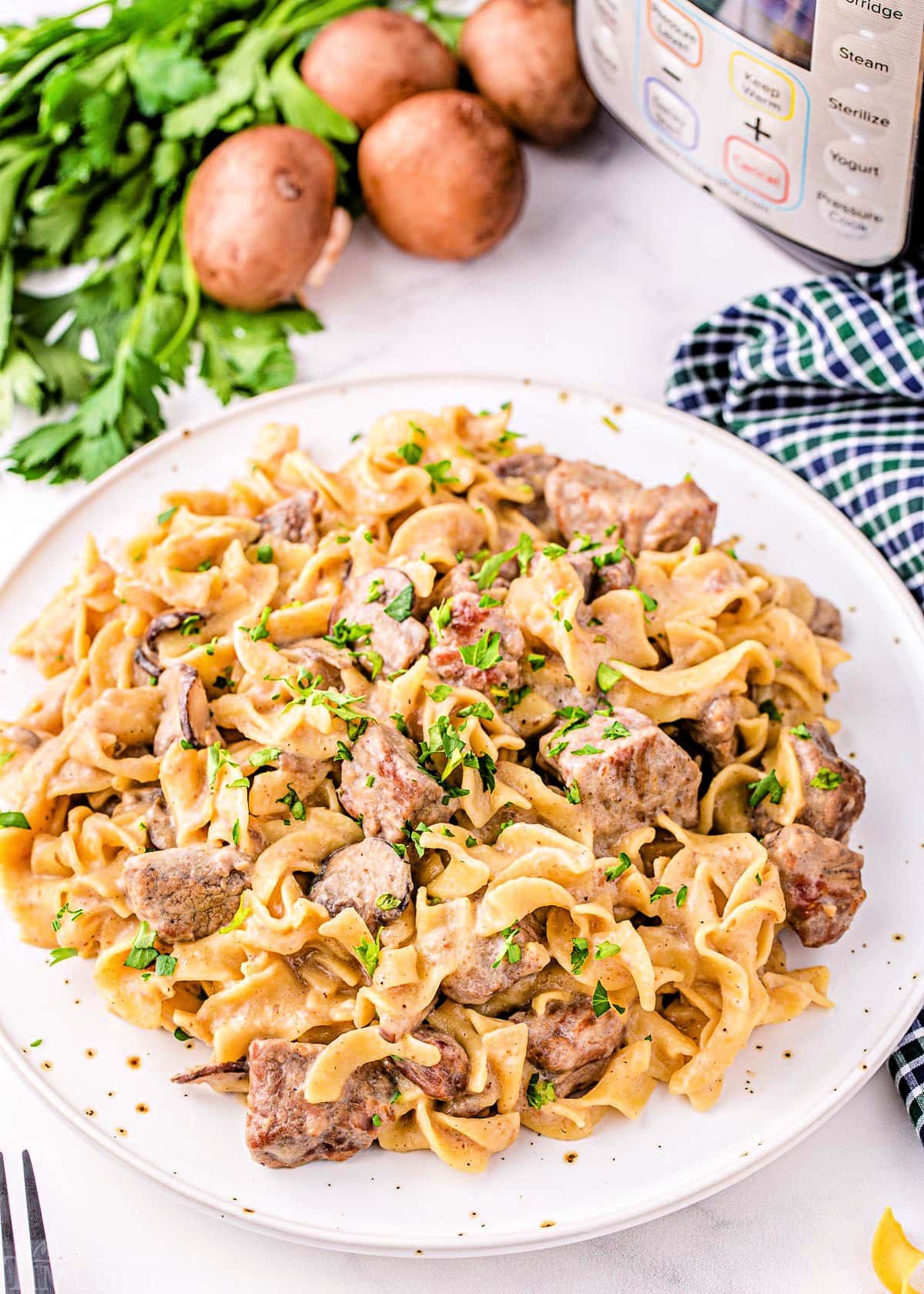 top down look at a white plate filled with beef stroganoff ready to be enjoyed. parsley and mushrooms are in the background next to the instant pot.