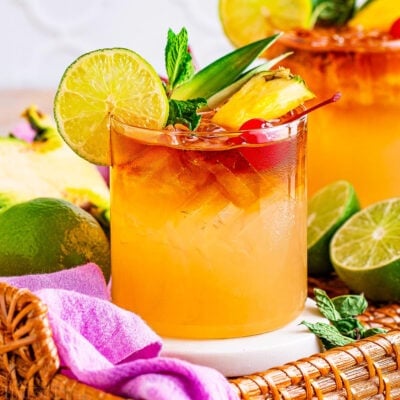 two mai tai cocktails prepared and garnished with lime, pineapple, maraschino cherries and are sitting on a rattan tray with a violet cloth napkin and cut limes around them.