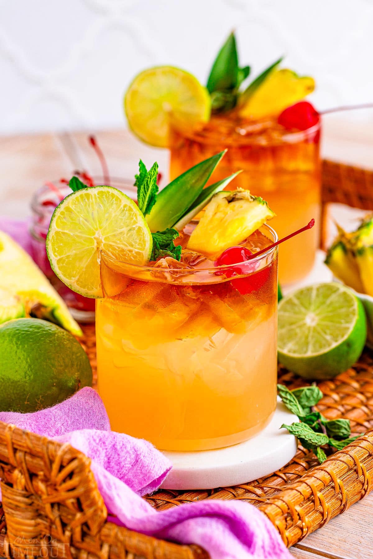 two mai tai cocktails prepared and garnished with lime, pineapple, maraschino cherries and are sitting on a rattan tray with a violet cloth napkin and cut limes around them.