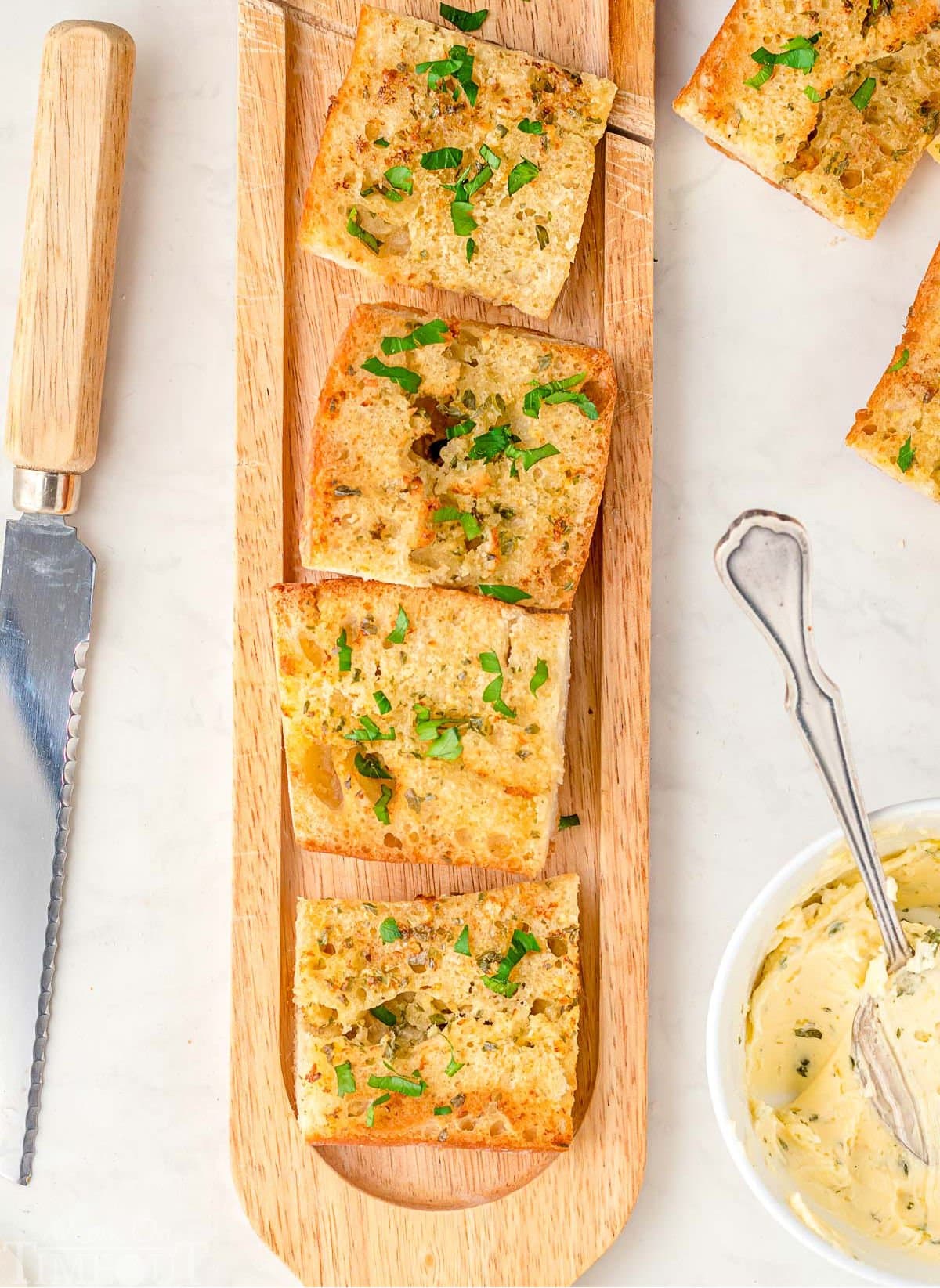 wood board with garlic bread cut into pieces on it. bowl of garlic butter off to the side with more pieces of garlic bread and a knife on the other side.