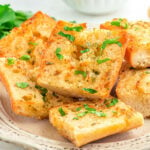 shot of garlic bread sliced and ready to serve off a white plate. fresh parsley garnish on the bread.