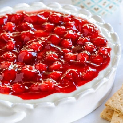 cheesecake dip topped with a can of cherry pie filling in a white scalloped pie plate.
