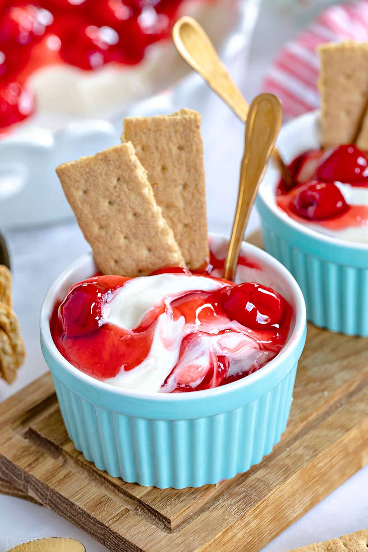 small aqua colored ramekin filled with cheesecake dip and topped with cherry pie filling. Two graham cracker pieces stuck into the dip along with a small spoon. The rest of the dip can be seen in the background.