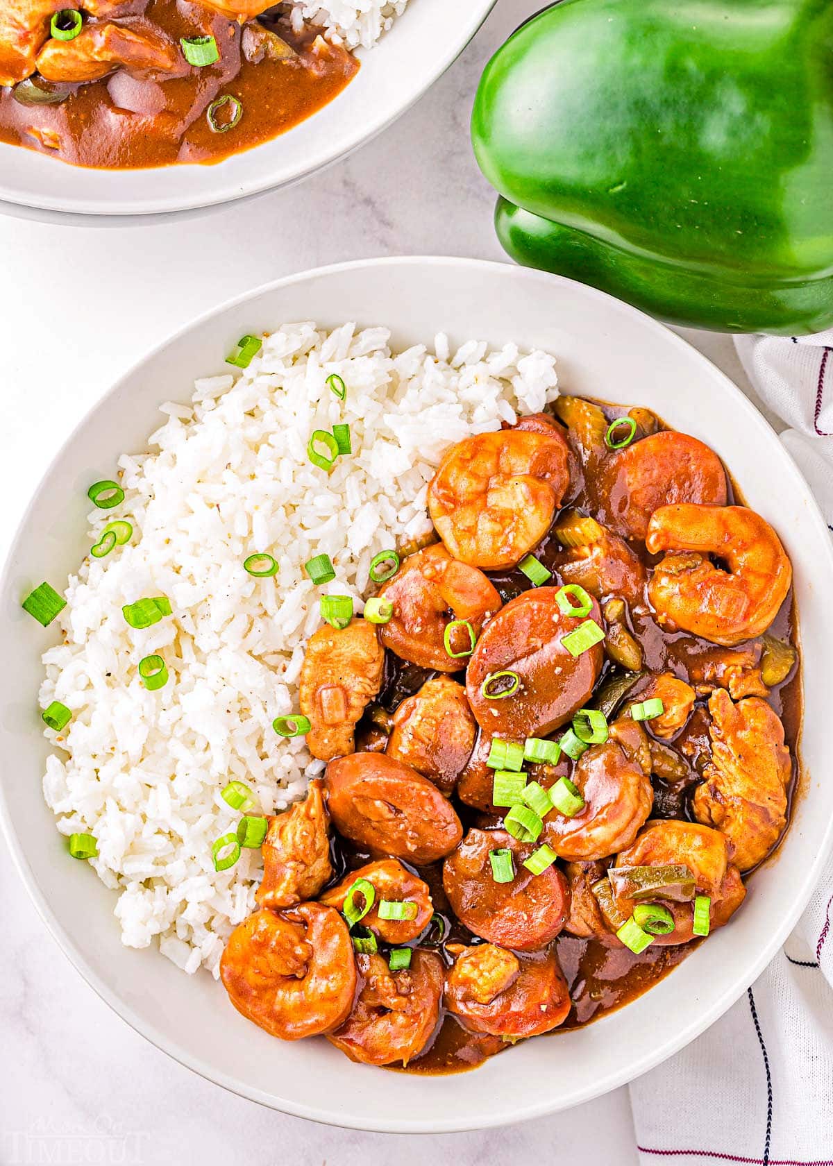 top down view of seafood gumbo served on a white plate with white rice and topped with green onions. part of another plate and a green pepper can be seen.