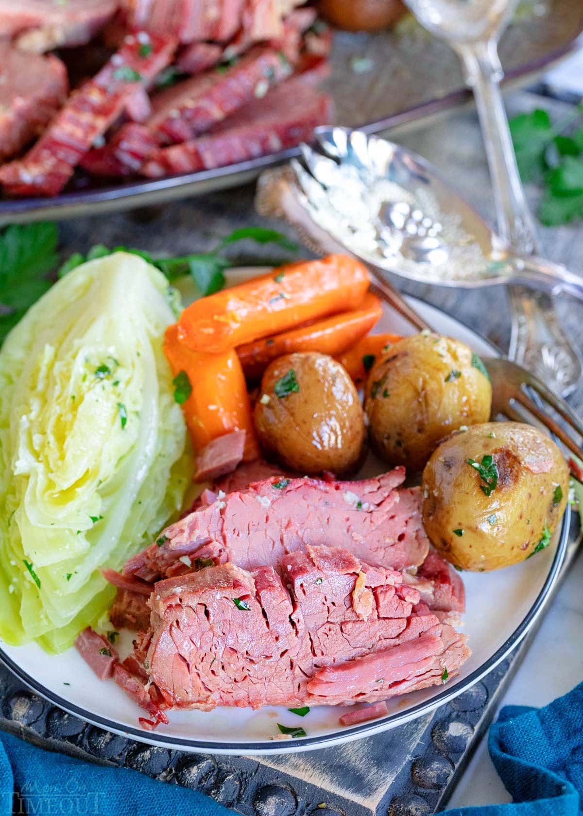 plate with a few slices of corned beef and cabbage, potatoes and carrots sitting on a gray wood board with serving plate in background.