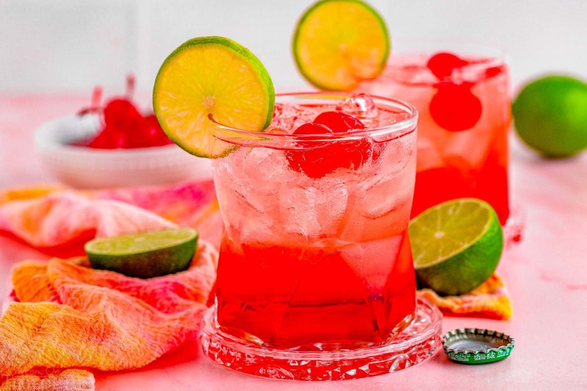 dirty shirley drinks in two short glasses. garnished with a lime slice on the rim and maraschino cherries in the drinks, additional garnishes sitting on the pink marble surface around the drinks.
