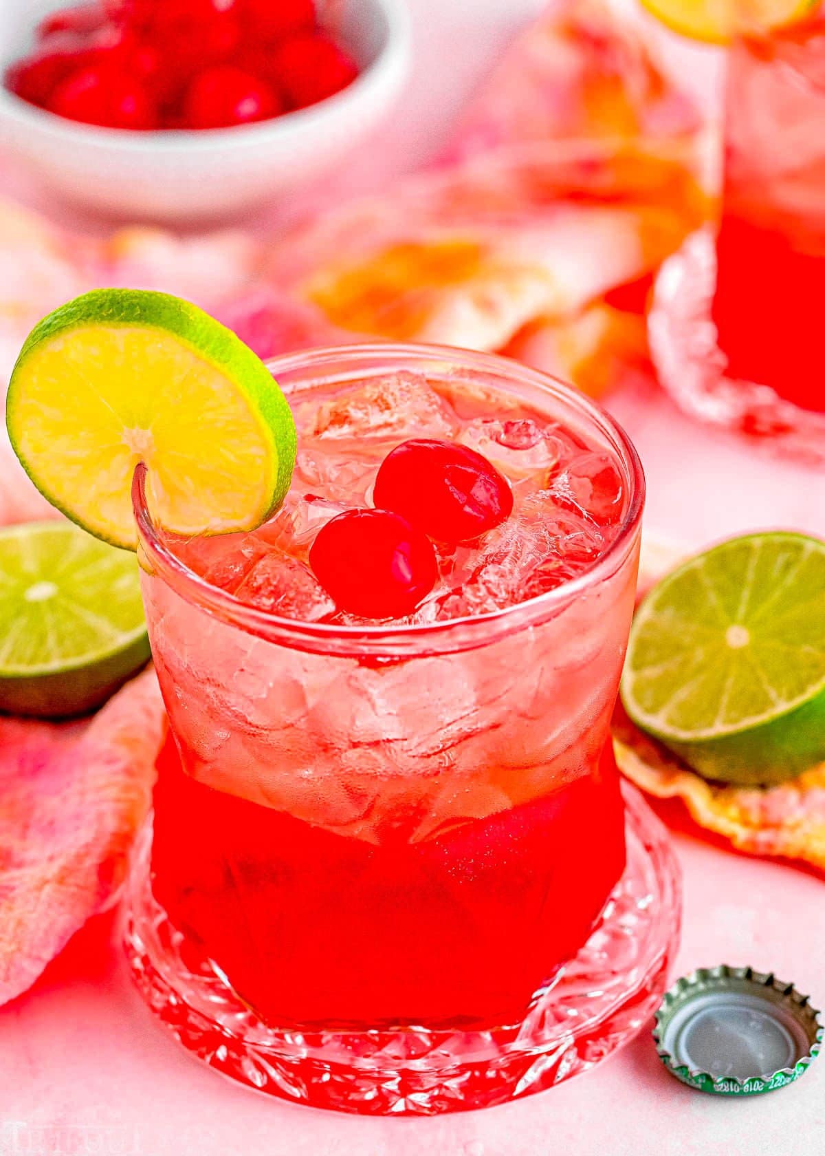 dirty shirley temple cocktail in a short glass garnished with a slice of lime and two maraschino cherries. Another drink and additional garnishes can be seen in the background.
