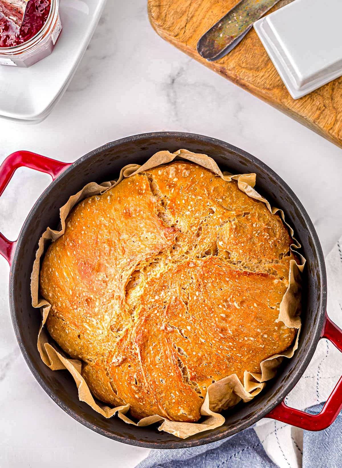 top down look at no knead bread in red dutch oven lined with parchment paper on a marble board. cutting board and jam are next to the dutch oven.