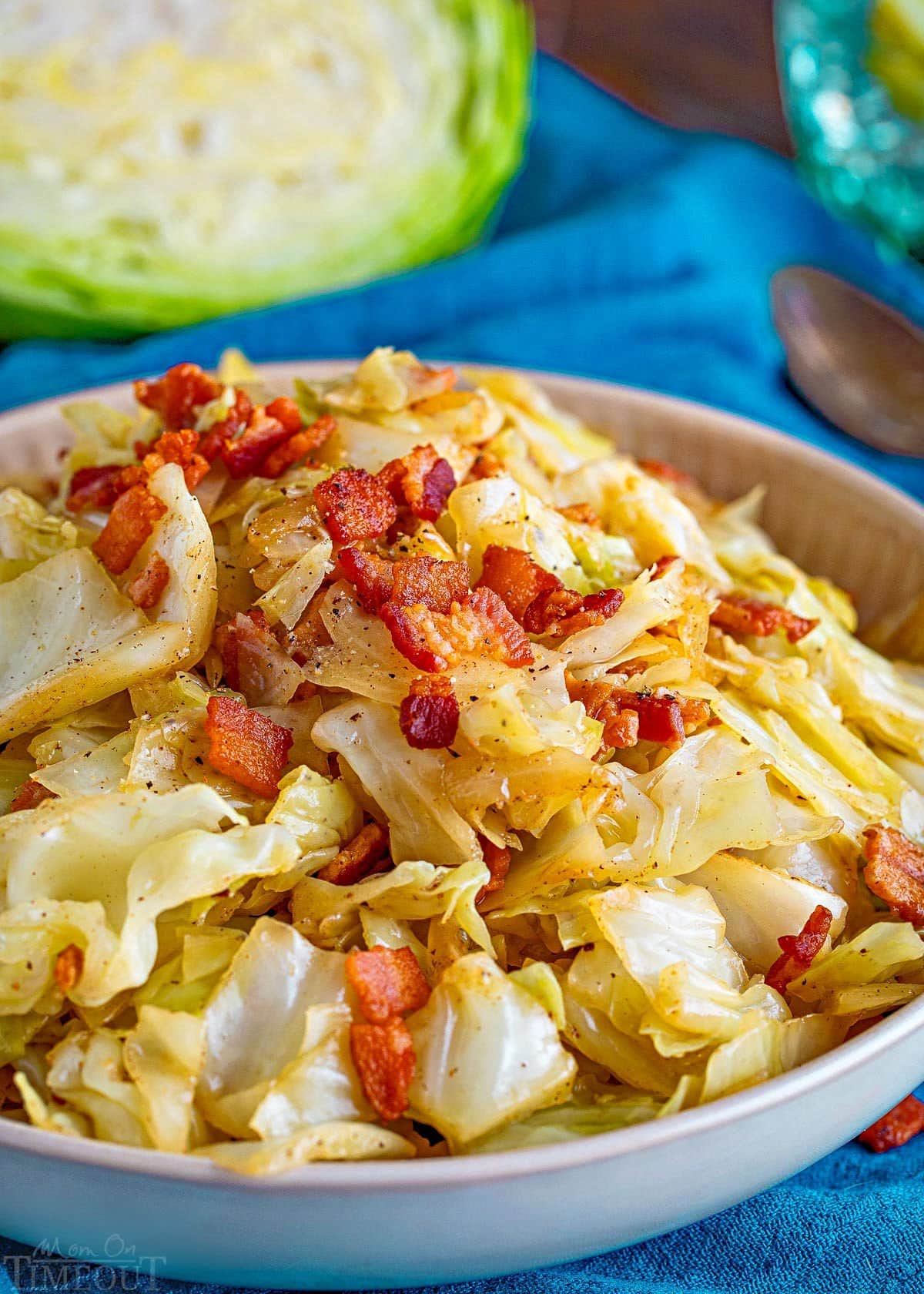 shallow bowl filled with fried cabbage made with bacon, onion and garlic. The bowl is sitting on a bright teal napkin and half a head of cabbage is in the background.