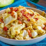 shallow bowl filled with fried cabbage made with bacon, onion and garlic. The bowl is sitting on a bright teal napkin and half a head of cabbage is in the background.