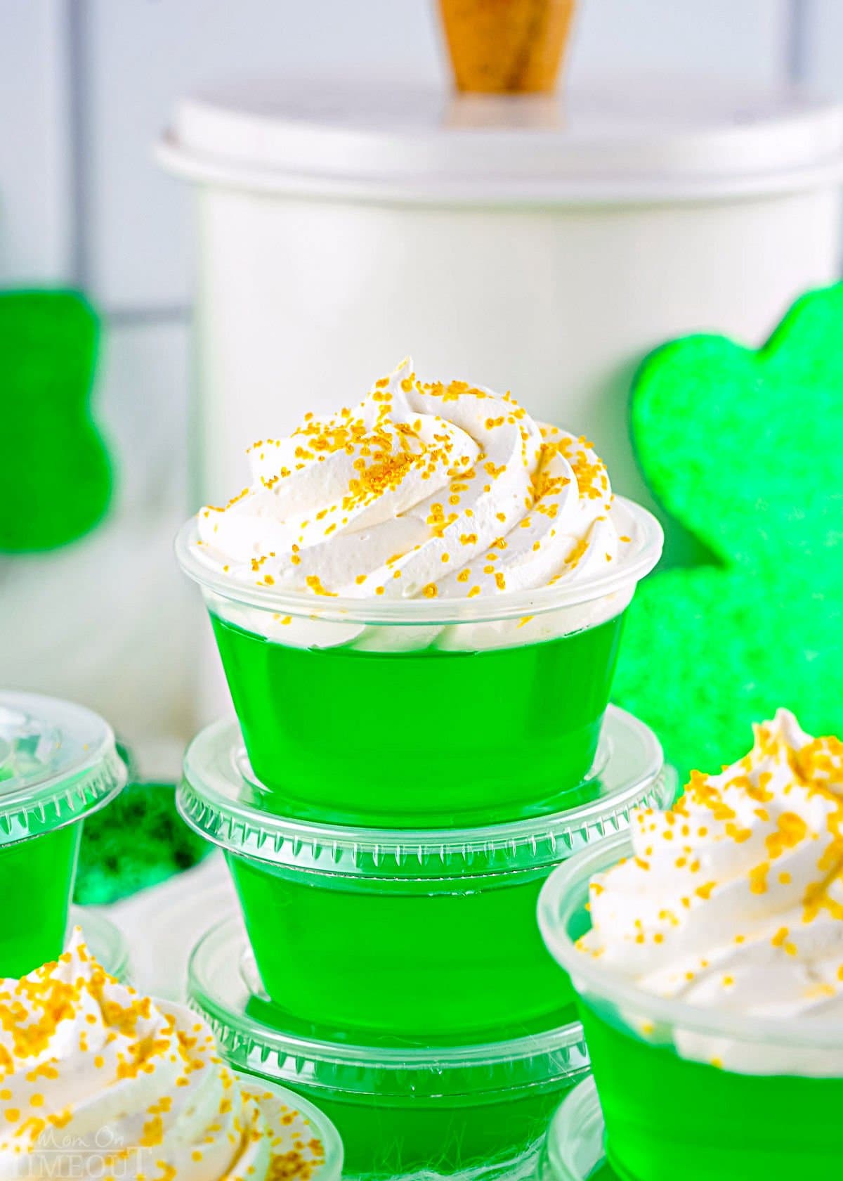 four green jello shots in small plastic cups topped with whipped cream and gold sanding sugar stacked on top of each other. white canister in the background as well as some st patricks day decor.