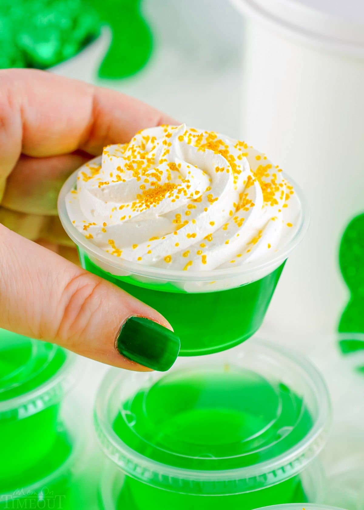 hand holding a green jello shot in front of more jello shots. the one that is being held has whipped cream on it.