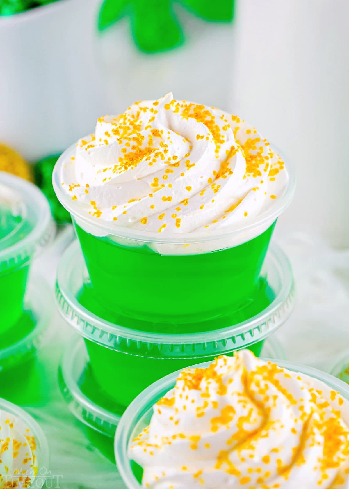 green jello shots in small plastic cups topped with whipped cream and gold sanding sugar stacked on top of each other.