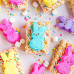 rice krispie treats decorated with white candy melts and sprinkles and topped with different colored Peeps. This is a top down view of the treats laid out on parchment paper.