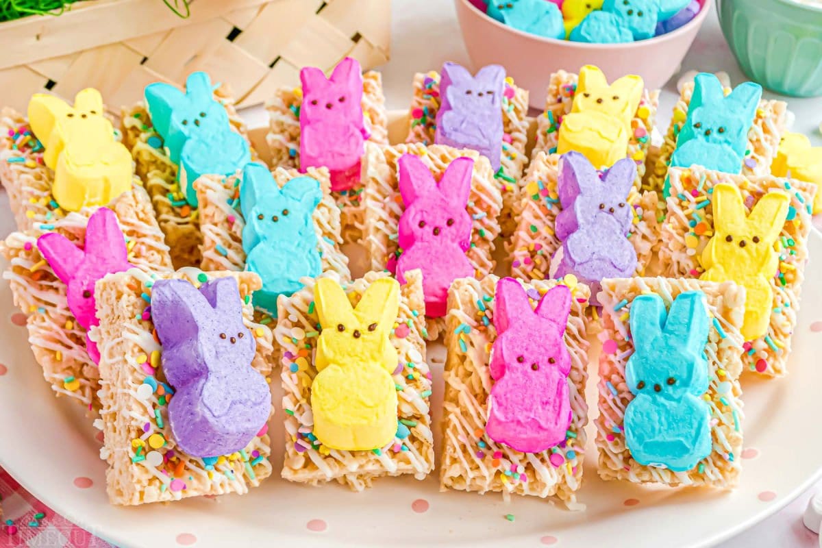 lots of rice krispie treats on a white plate topped with different colored peeps ready to be enjoyed.