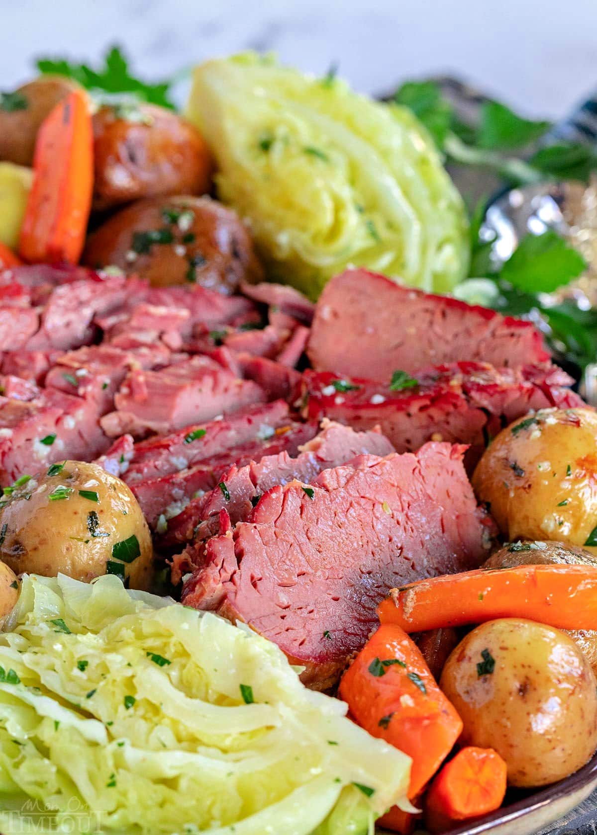 close up look at slices of slow cooker corned beef and cabbage wedges plus potatoes and carrots on a serving platter brushed with garlic herb butter.