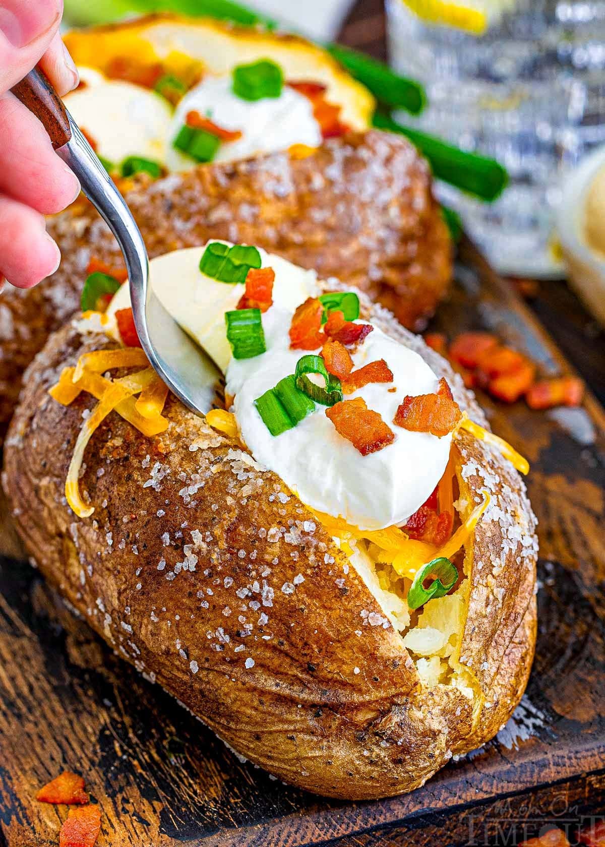 fork is inserted into the loaded baked potato as if ready to pull out a bite. potatoes are sitting on a dark wood board.