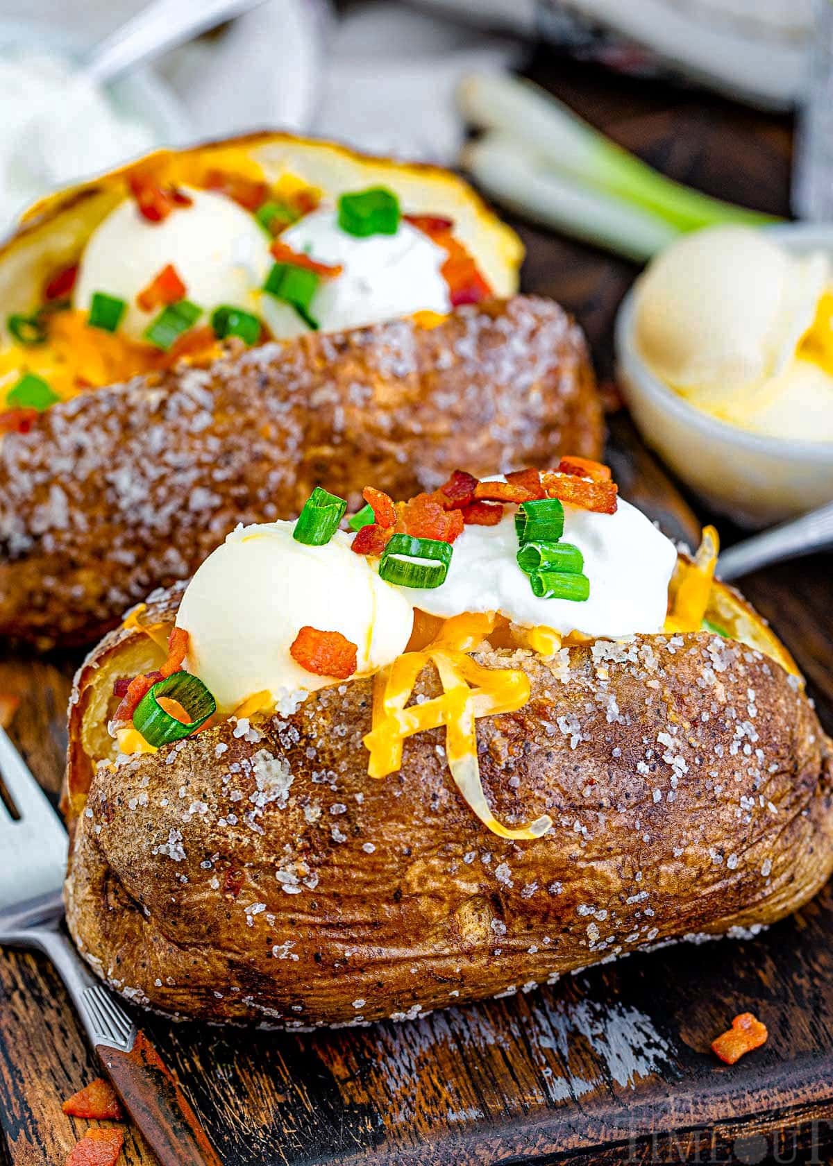 angled look down at two baked potatoes topped with cheese and sour cream and bacon ready to be enjoyed. You can see the salt on the skin of the potato.