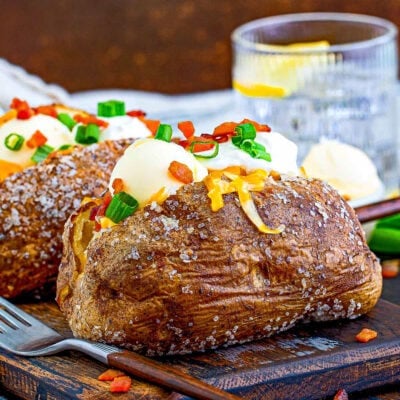 two loaded baked potatoes sitting on a dark wood board topped with sour cream, butter, bacon, green onions and cheese. glass of water in background.