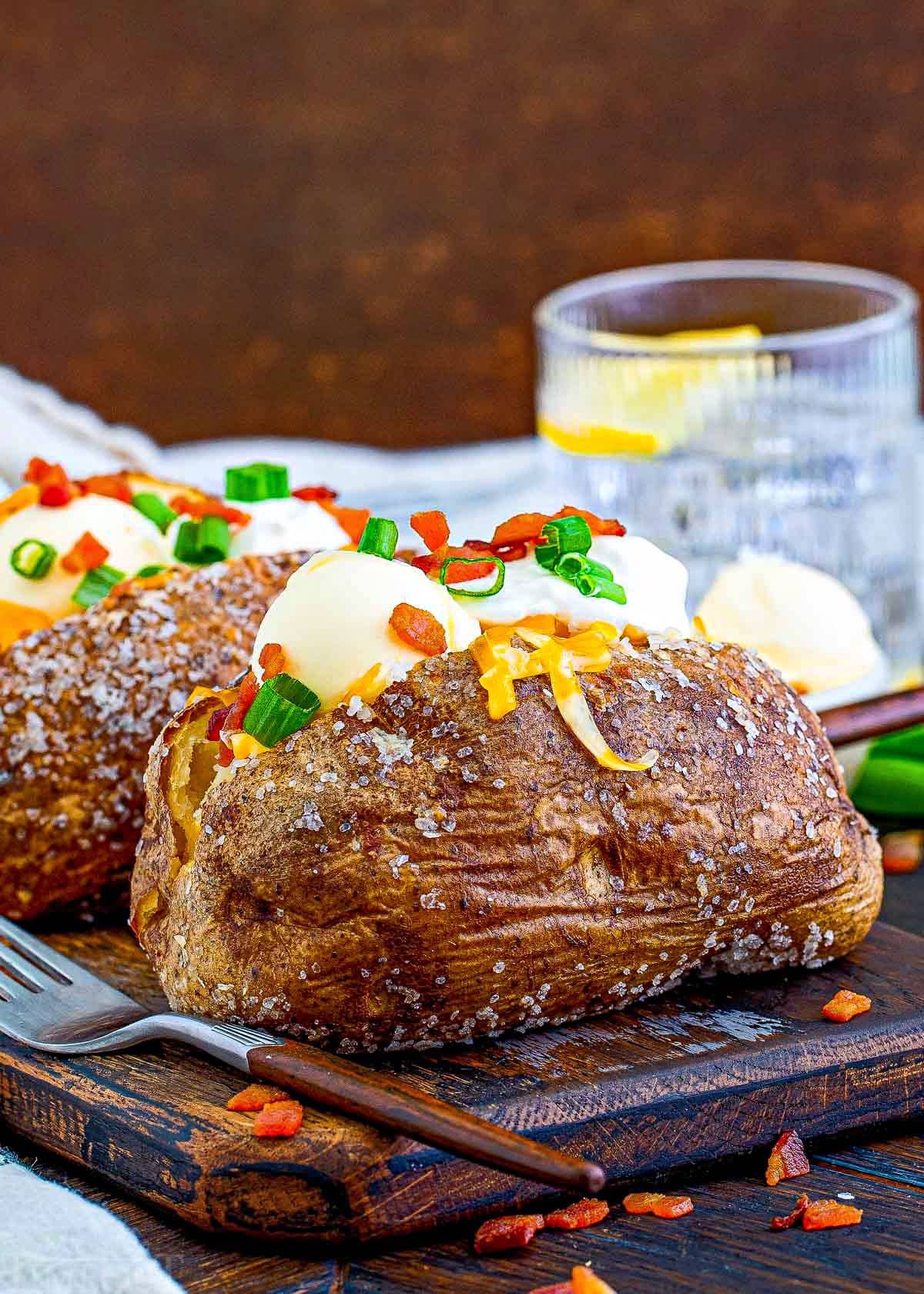 two loaded baked potatoes sitting on a dark wood board topped with sour cream, butter, bacon, green onions and cheese. glass of water in background.