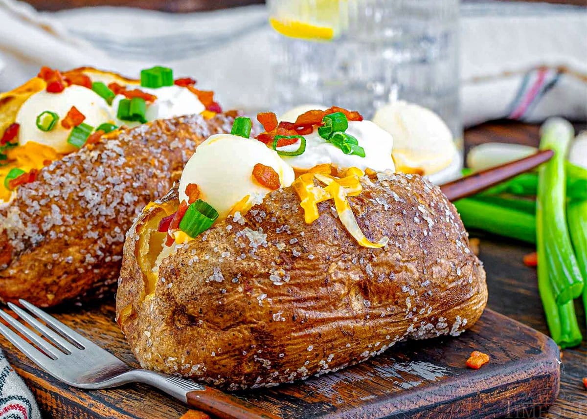 two loaded baked potatoes sitting on a dark wood board topped with sour cream, butter, bacon, green onions and cheese. glass of water in background.