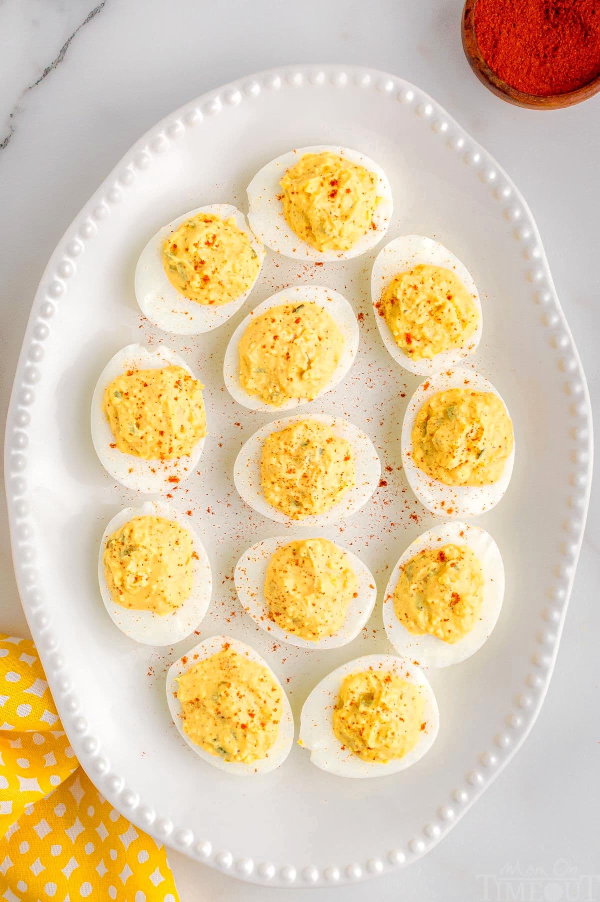rimmed white serving tray with 12 deviled eggs on it. Eggs have been garnished with paprika and there is a yellow napkin in the corner.