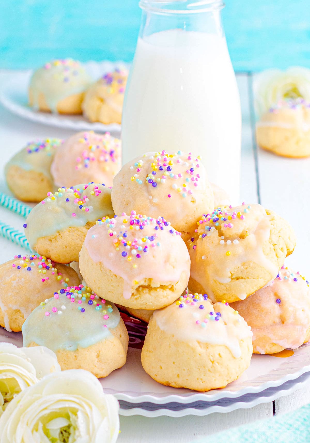 plate of Italian Cookies sitting on front of a jug of milk and decorated with a pastel glaze and topped with sprinkles. Aqua background and more cookies can be seen in the background.