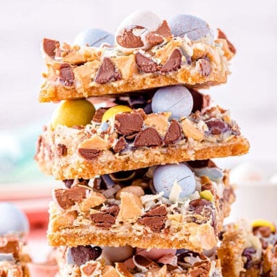 magic cookie bars topped with cadbury easter eggs stacked four high on a white surface.