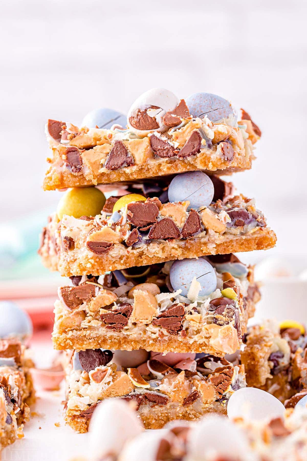 magic cookie bars topped with cadbury easter eggs stacked four high on a white surface.