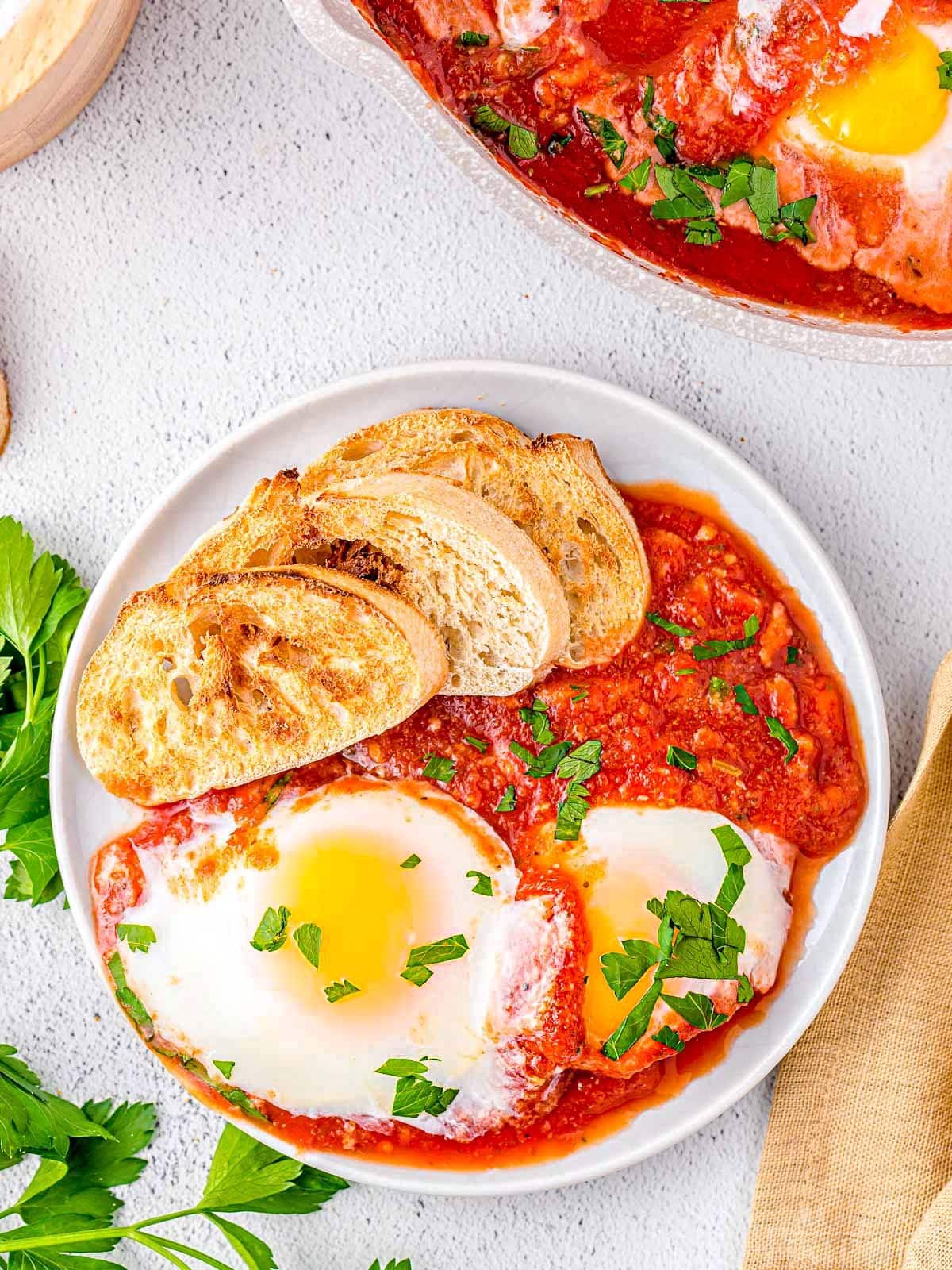 top down look at plated eggs in purgatory with two perfectly poached eggs rested in tomato sauce. Crusty, toasted bread on the plate. Garnished with chopped parsley.