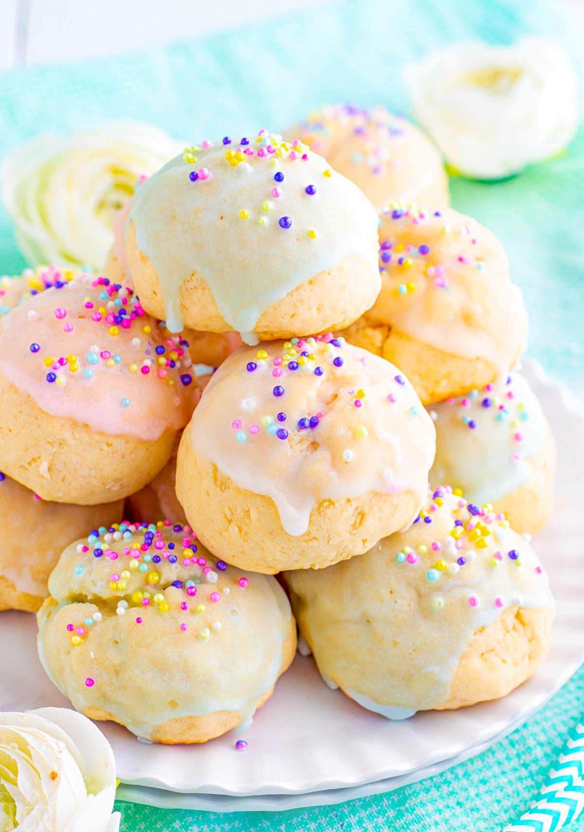 coloful italian cookies piled high on a white scalloped plate. cookies are topped with sprinkles and pastel glaze in varying colors.