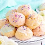 wide view of colorful italian cookies piled high on a white scalloped plate. cookies are topped with sprinkles and pastel glaze in varying colors.