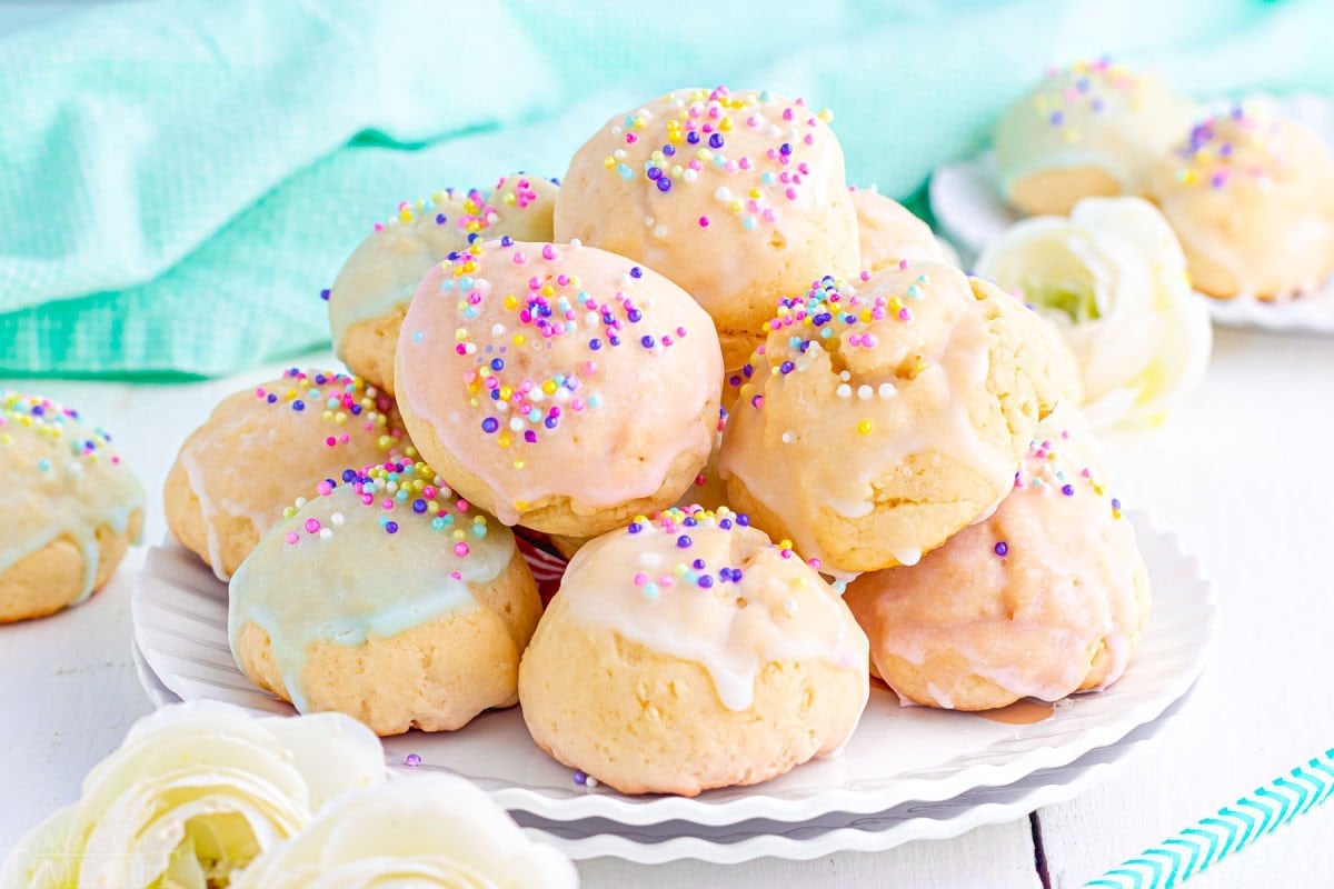 wide view of colorful italian cookies piled high on a white scalloped plate. cookies are topped with sprinkles and pastel glaze in varying colors.