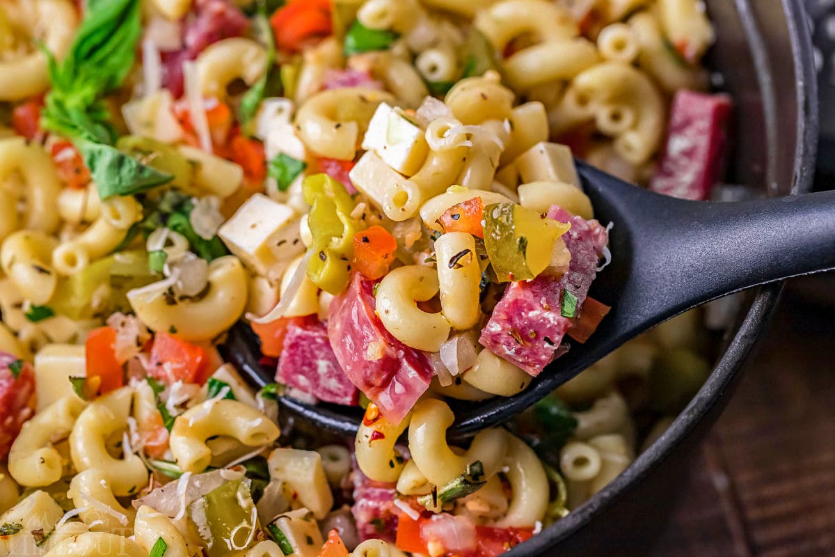 serving spoon inserted into the pasta salad made with italian dressing, salami, pickles and more.