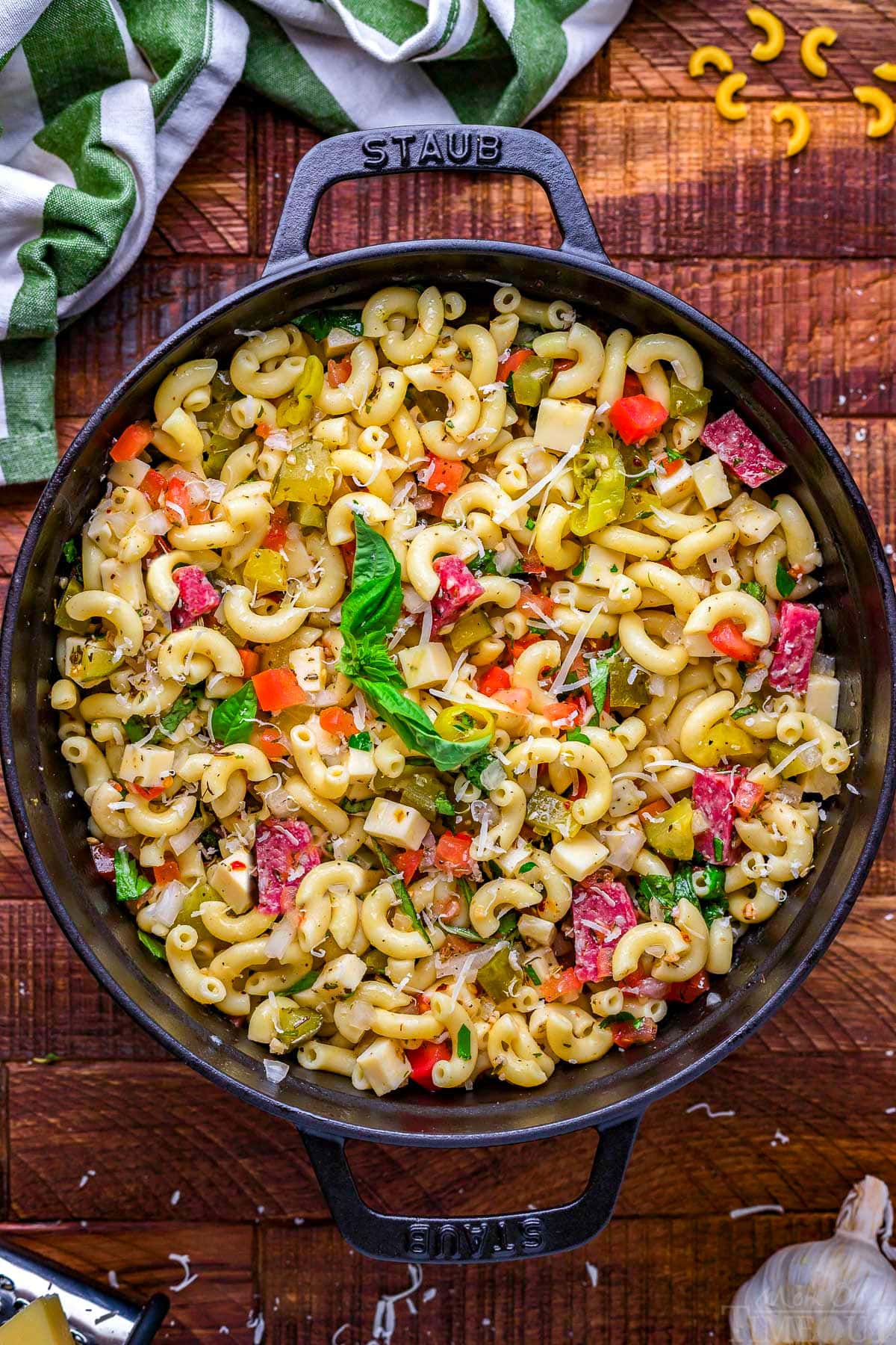 top down look at italian pasta salad ready to be served out of a black staub round dish with handles. dark wood surface beneath the pot.