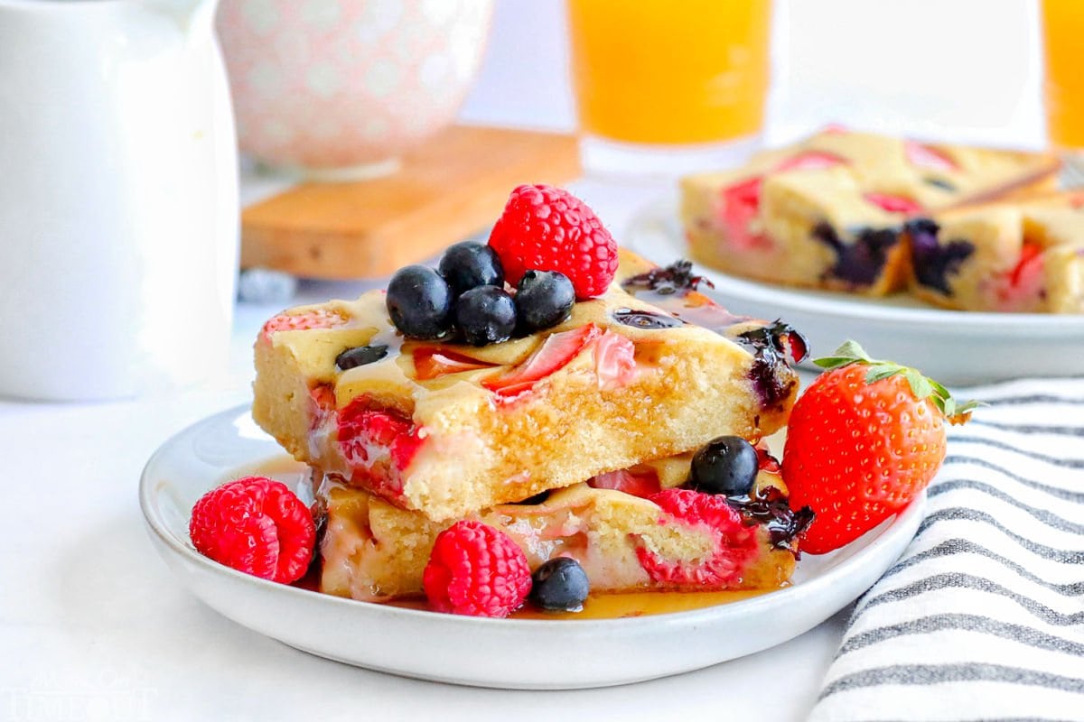 two sheet pan pancakes cut into squares stacked on a white plate and topped with with fresh berries and maple syrup.