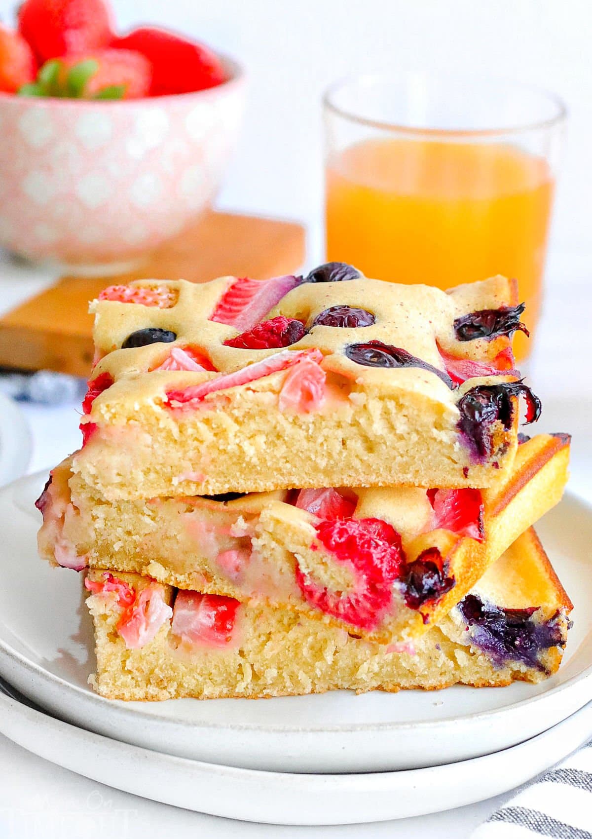 three square berry pancakes stacked on a two white plates with a glass of orange juice in the background.