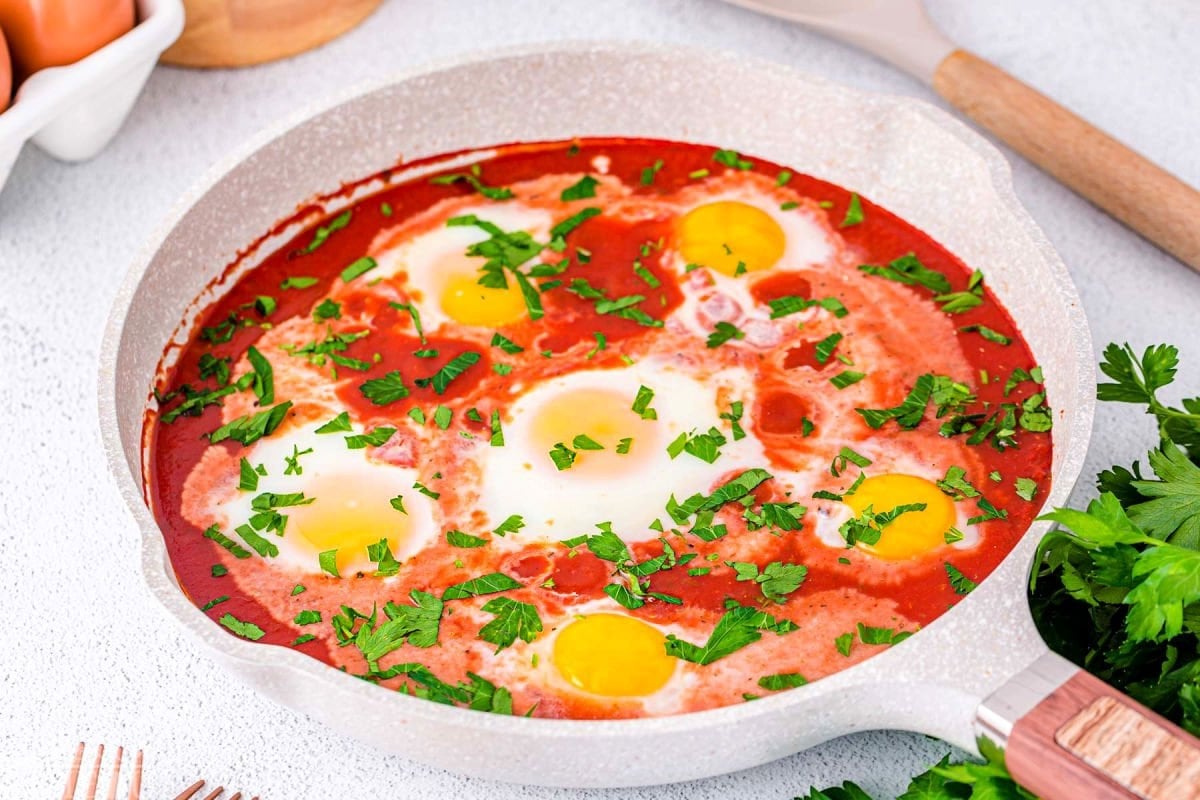 wide shot of light colored skillet with six eggs poached in tomato sacue and topped with fresh parsley.
