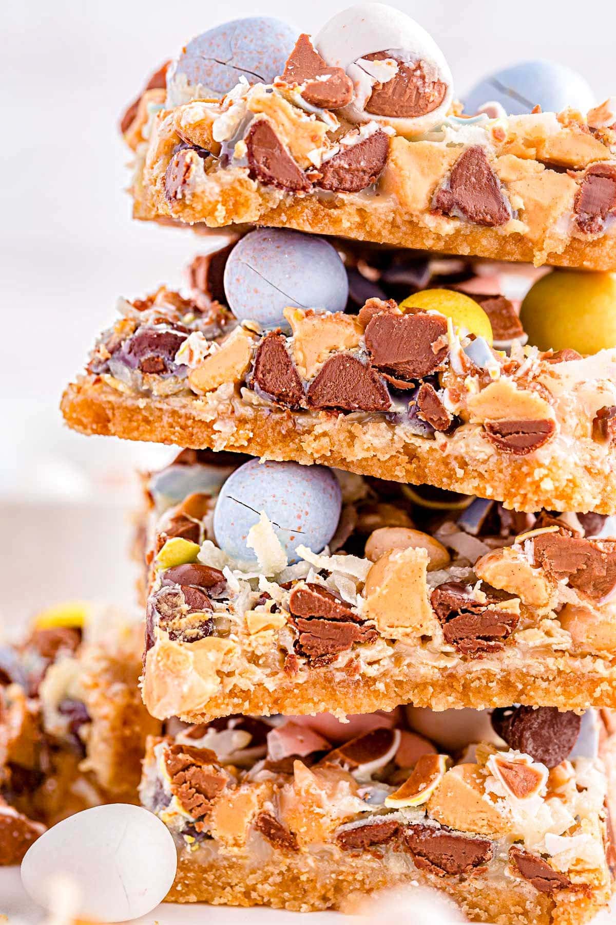 close up look at magic cookie bars topped with cadbury easter eggs stacked four high on a white surface.