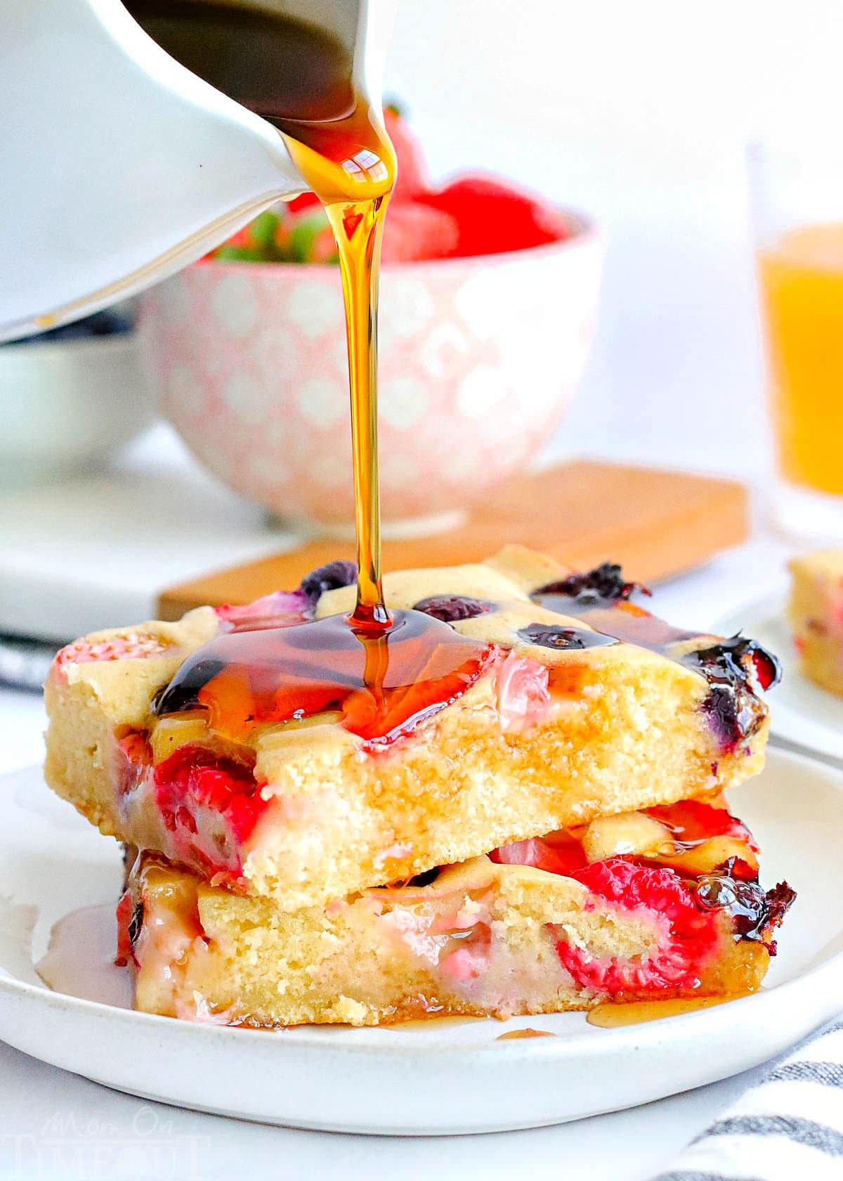 two pieces of sheet pan pancakes made with three types of berries stacked on a white plate. syrup is being poured on top of the pancakes and a bowl of berries can be seen in the background.