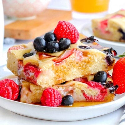 two sheet pan pancakes cut into squares stacked on a white plate and topped with with fresh berries and maple syrup.