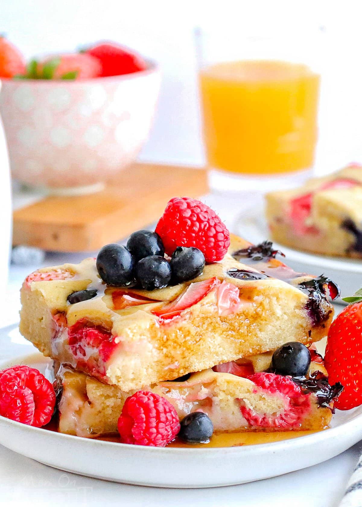 two sheet pan pancakes cut into squares stacked on a white plate and topped with with fresh berries and maple syrup.