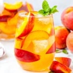 peach sangria recipe made with white wine in stemless wine glass ready to be enjoyed. garnished with mint and fresh peach slices.